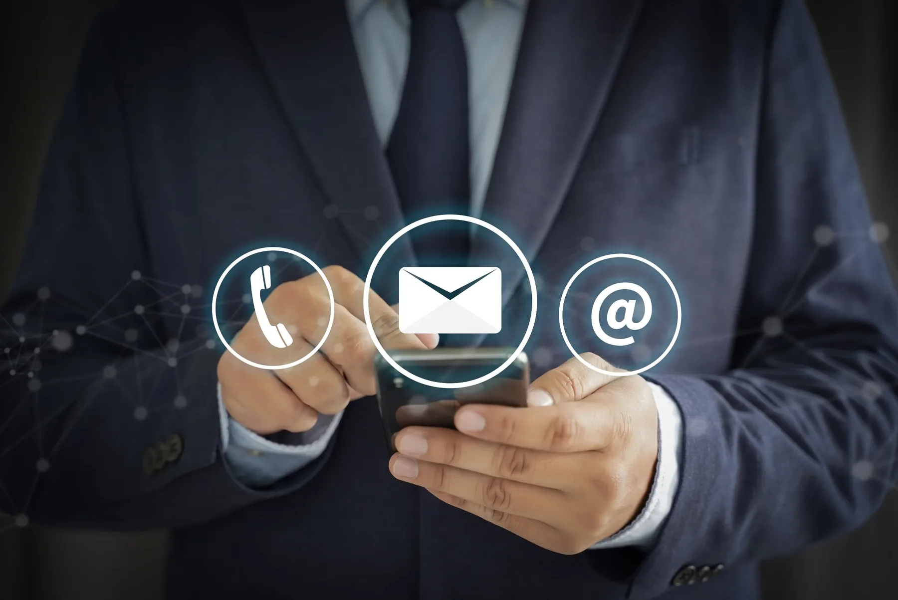 A person in a dark suit holding a smartphone, with phone, envelope and at-sign icons displayed above it.