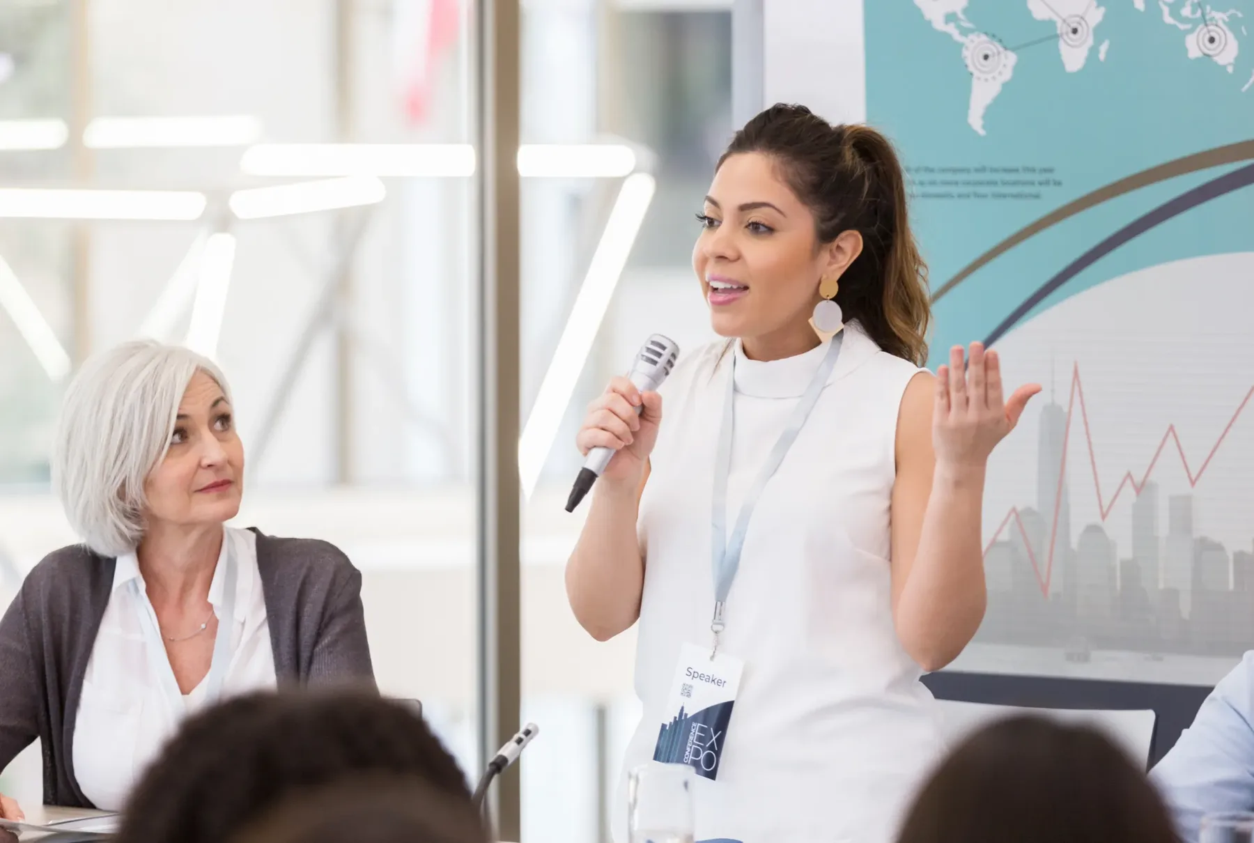 A woman speaking to a room with a microphone in her hand while a colleague listens beside her at a conference panel.